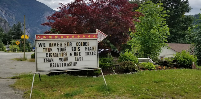 A sign in front of a lush green mountainside reads "We have beer colder than your ex's heart. Cigarettes more toxic than your last relationship."