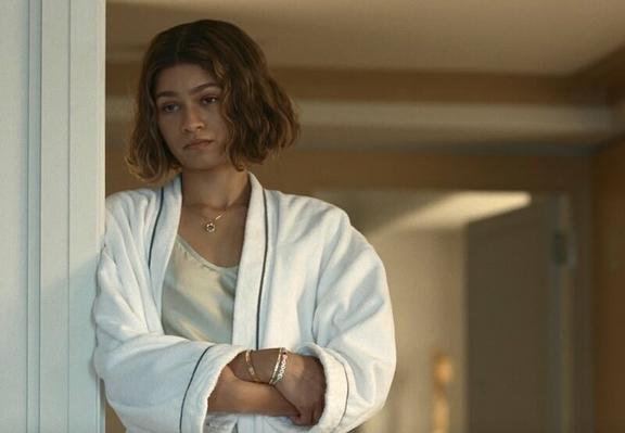 Zendaya wears a white bath robe. She leans against a door jam. She's looking at someone seated near her.