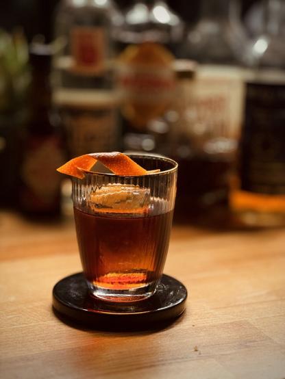 rich red brown cocktail with a big rock of clear ice and orange twist garnish sitting on a black ceramic coaster in front of bottles of Bitter Truth chocolate bitter, Alipús mezcal, Amaro Averna, Skipper’s Finest demerara rum, Smith and Cross jamaican rum, and a maple syrup bottle in the shape of a maple leaf