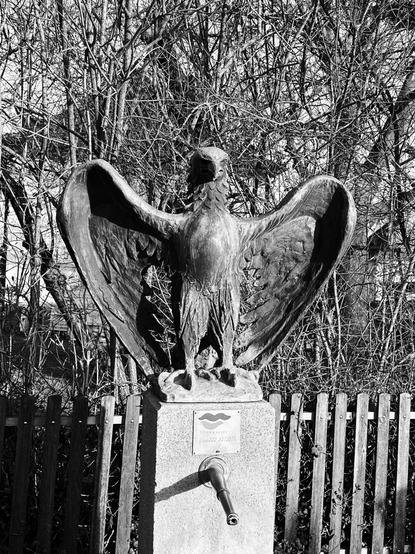 Eine Steinadler-Statue mit ausgebreiteten Flügeln steht auf einem Sockel vor einem Holzzaun und Bäumen im Hintergrund.