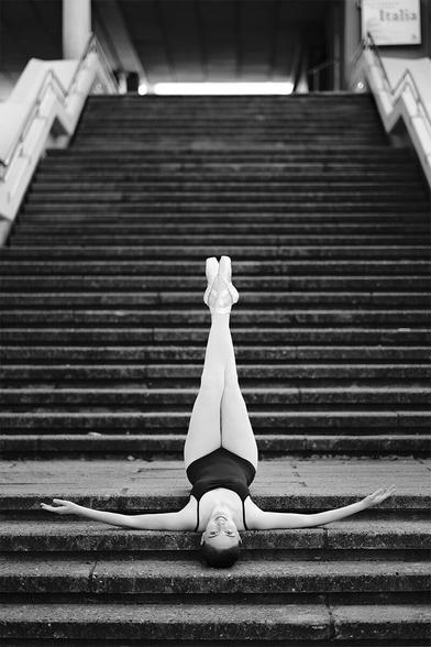 Ballerina, die mit Kopf nach unten rücklings auf einer Treppe liegt, die Arme ausgestreckt und die Beine nach oben gestreckt hat.