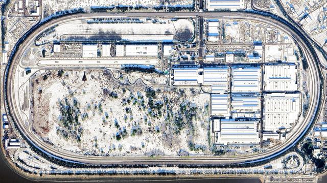 A satellite picture of an industrial landscape, taken during the winter. All the landscape is mostly white due to snow. A large oval automotive circuit is surrounding the assembly plant.
