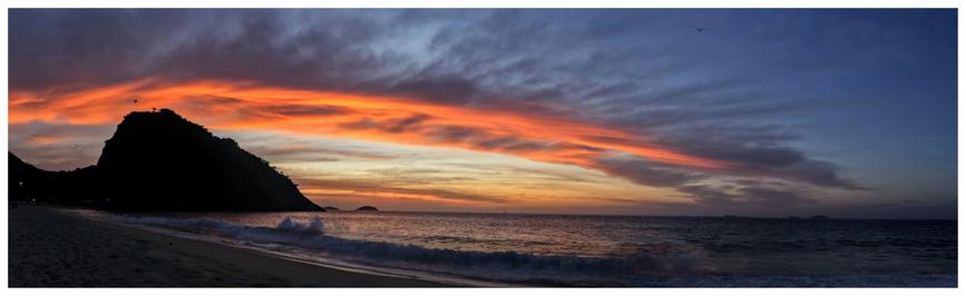 A panorama photo of a colorful sky seen before sunrise on the beach front at "praia do Leme", in Rio. The clouds are orange and the sky colors range from blue to magenta tones.