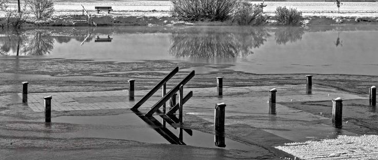 Schwarzweißfotografie im Format 21:9 waagerecht.
Winter an einem Badesee. Es liegt wenig Schnee, ist aber nicht kalt genug für eine geschlossene Eisdecke.
Im Vordergrund ein Badesteg mit Treppe. Das Wasser steht so hoch, dass die Steegfläche gerade unter Wasser ist und die Handläufe der beiden Treppengeländer aus dem Wasser heraus ragen. Das gegenüberliegende Ufer ist als schmaler Streifen zu erkennen. Dort, links oben, führt eine Treppe aus dem Wasser, neben welcher rechts eine Sitzbank aus Holz steht.