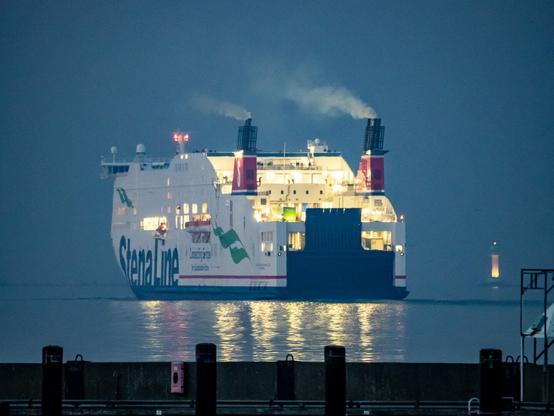 Das Fährschiff Stena Scandinavica verlässt hell erleuchtet den Hafen. Man sieht das Heck der Fähre mit der großen dunklen Klappe, das Licht spiegelt sich auf dem Wasser, aus den Schornsteinen kommt Dampf, im Vordergrund ist eine Kaimauer des Yachthafens zu sehen, rechts im Hintergrund der Leuchtturm Falckenstein.