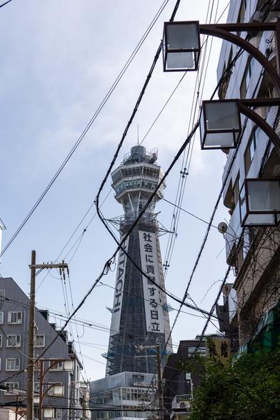 Tsutenkaku, a landmark tower in Osaka looms behind some crisscrossed wires hanging over the street from where the picture is taken. The tower is wrapped in scaffolding as it was undergoing refurbishment at the time.