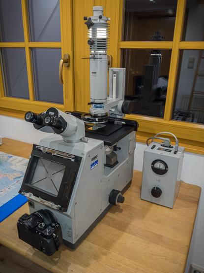 An old ZEISS ICM405 inverted microscope. It's pretty big and has the typical old hardware grey color. Since it's inverted, the objective revolver sits under the specimen stage and looks up. In the front it has a binocular for viewing, and a DSLR is attached to the camera port below. A large variable transformer sits next to the device for regulation of the light bulb.