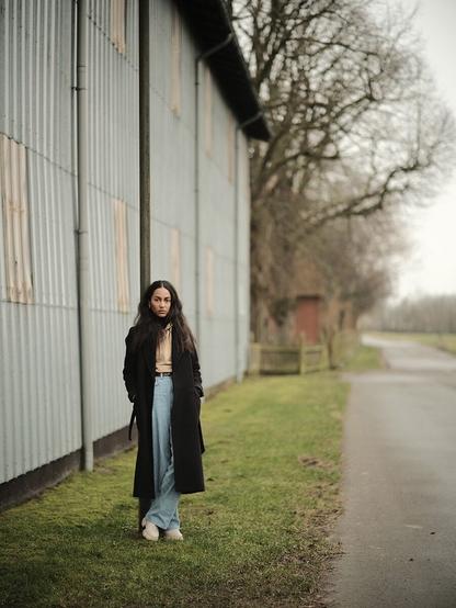 Woman standing on the side of a rural road in front of a barn leaning on a street lamp, looking into the camera