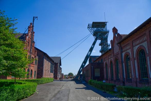 A photo of the Zeche Zollern. On the left and right you can see red brick buildings. On the left is the administrative building and machine hall. On the right the Lohnhalle, Schwarzkaue and Lampenstube. There are green hedges and tree's on the left. Behind the building on the right is a winding tower. The sky is clear blue.