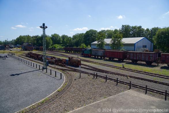 A view out of the mine hall towards a railyard with old trains and wagons. Mostly rusty and damaged. Beheind the railyard are green trees. The sky is clear blue with some light white clouds.