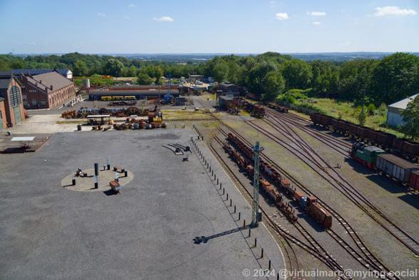 A photo from the winding tower over the trainyard and the place in front of the machine hall of Zeche Zollern. Theres green landscape with many trees around the area of the coal mine. The sky is clear blue.