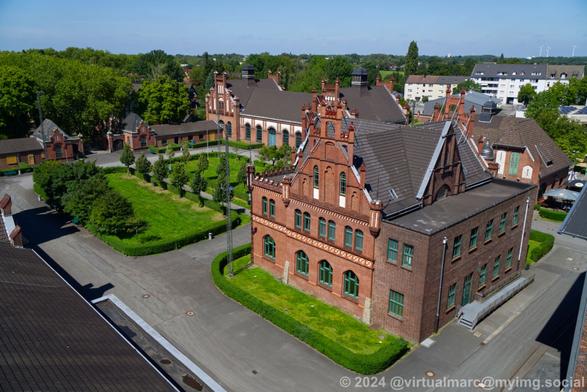 A photo from the winding tower over the entrance and main place of Zeche Zollern. You can see the entrance gate with many trees on the way outside, the entrance buildings, the grass area sourrounded by hedges and containing trees in front of the administrative building and the administrative building sourrounded by grass and hedges. The sky is clear blue.
