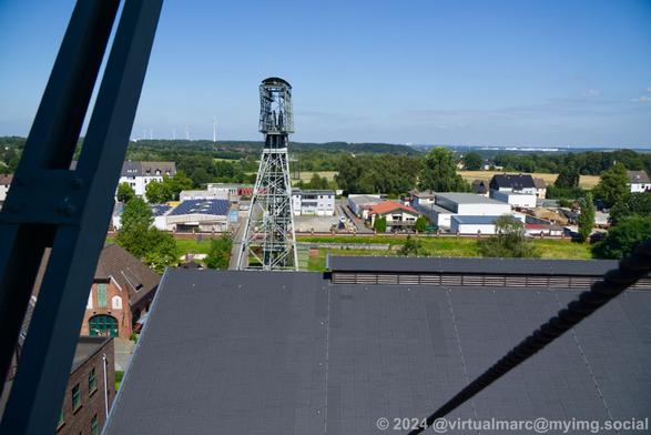A photo from the winding tower towards the left winding tower and the area beyond with a small industrial area and many trees and farmland. You can see some wind turbines in the distance. The sky is clear blue.