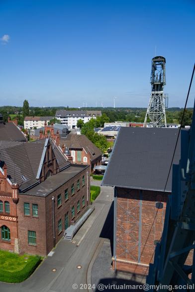 A photo from the winding tower to the left winding tower. You can see part of the machine hall and the administrative building and the green landscape with the wind turbines.