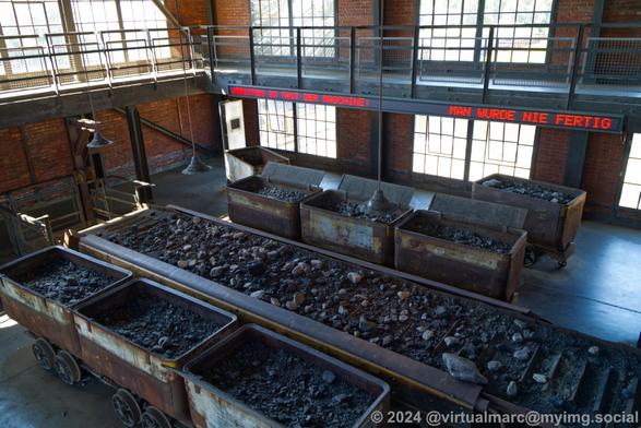A view from the top level of the mine hall towards the bottom level. There are multiple minecarts with coal in it. A conveyor belt with coal and stones on it is going through the room. Theres a rolling text on a wall which reads: "Arbeiten im Takt der Maschine: Man wurde nie fertig" (Translation: "Working in time with the machine: You never finished")