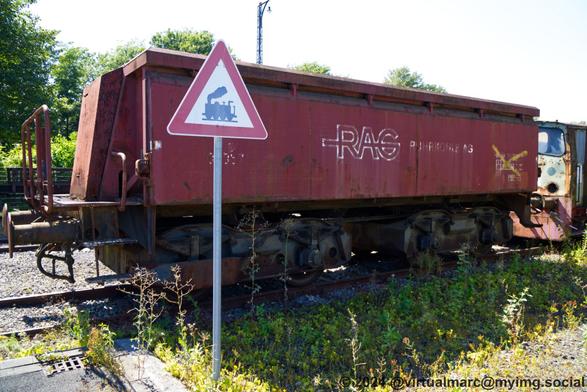 A photo of a railway wagon in Red with the Logo of RAG (Ruhrkohle AG) on rail tracks behind a warning sign for crossing trains.