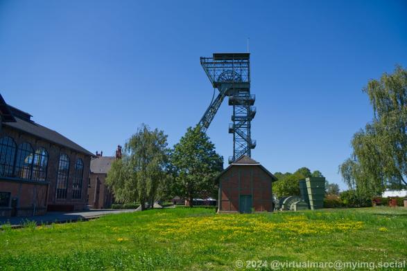 A photo of a small shed made out of red bricks with green accents on a green grass. Part of the machine hall with big windows can be seen on the left. Behind the shed and some Trees you can see the left winding tower.