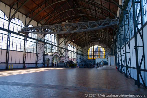 A photo of the inside of the inside of the machine hall with a big empty space in the front. You can see machines in the distance. There is a big industrial crane bellow the ceiling. You can see big windows on all sides of the building.
