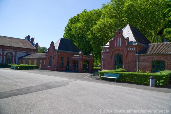 A photo of red brick buildings which are the entrance buildings and doors of the area. Some green hedges with benches are in front of the entry/exit buildings.