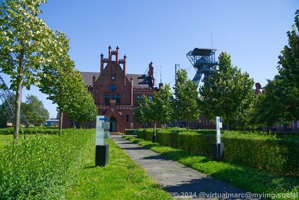 A view to the administrative building through the entrance area between two grass areas sourrounded by hedges with trees in them. You can see the right winding tower on the right of the administrative building behind some trees.