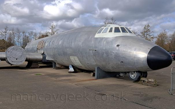 Side view of a rather dilapidated, bare metal, 4 propellor-engined airliner (with the engines removed!) lying on blocks on the floor.
The nose-wheel is slightly dropped down, and resting on another block.
The metal fuselage appears to be torn around the wing root, probably where the wing was removed to transport it here.
The frame around the cockpit windows appears to have been painted white, which look a bit odd with the rest of it being in a bare metal finish.
Fluffy grey and white clouds fill most of the frame behind.