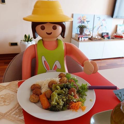 Mulher playmobil grande de camisola rosa e avental verde e chapéu largo amarelo sentada à mesa com um prato de legumes