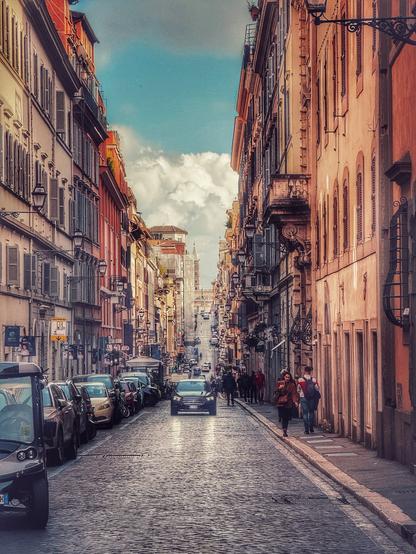 A long straight city street in Rome, with tall buildings on either side. There is a car in the middle of the road, heading towards the camera.
The sky is blue, but with large fluffy clouds
