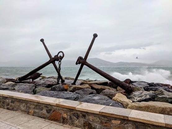 Two large, weathered, rusty anchors are positioned on a rocky breakwater beside a turbulent sea. The anchors lie horizontally, their flukes resting on the dark grey and brown rocks. On the anchor on the left, a heavy, rusted chain connects to the shank and extends towards the other anchor.
Large waves crash against the rocks, creating white foamy splashes. The sea itself is a choppy, grey-green under an overcast sky. In the distance, hazy, low-lying mountains can be seen across the water. A single bird is visible flying in the middle distance.
In the foreground, a paved walkway runs alongside the breakwater, bordered by a low wall constructed from rough-hewn stones. The overall scene conveys a sense of maritime history and the power of the sea on a blustery day.
