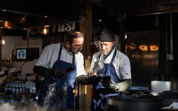Two chefs at a paella stall in Borough Market are talking to each other. Both wear black gloves and blue aprons. They are standing next to each other, one holds a phone and shows it to the other, they both look at the screen. Sunlight hits their faces while the background is rather dark