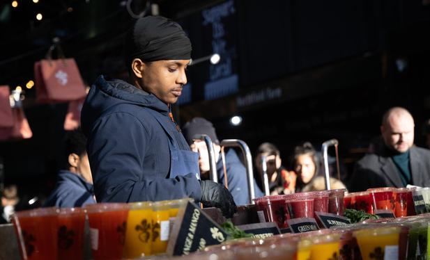 A vendor at a stall in Borough Market that sells smoothies. He is wearing a black apron and a black headscarf and is placed behind a row of ready made smoothies in clear plastic cups.