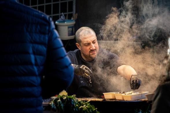 A closer shot of the chef from the previous foto, he leans forward to the pan and sprinkles cheese onto the food.