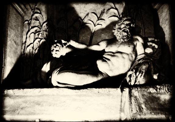 A monochrome picture of a Dionysus/Bacchus statue in Rome, above a street fountain. Night time and under lit casting dramatic shadows.