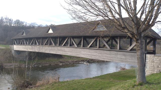 Eine Fachwerkholzbrücke über die begradigte Wutach. Das Dach der Brücke hat drei dreieckige Dachfenstergiebel.