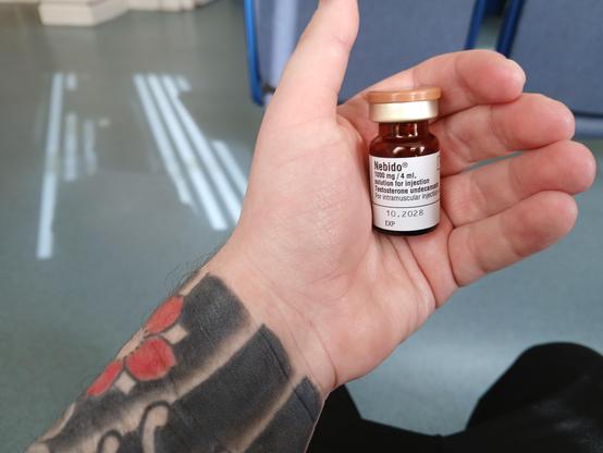 A white hand holding a vial of nebido. There are Japanese tattoos on what is visible of the forearm, and a nhs waiting room in the background