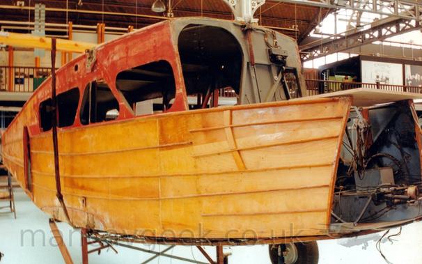 Side view of the bare wooden fuselage of a small, pre-World War 2-era airliner, standing on metal supports, facing to the right, looking more like a boat than an aeroplane.
Most of the exterior is yellow, with the upper sections being an orangey-red, and the interiors being painted a flat grey.
Mounting points for the bi-plane wings can be seen at the top and bottom of the fuselage.
There is a gaping hole where the front of the cockpit should be, allowing a view of wires and pulleys for the controls, as well as internal bracing struts.
The large, ovoid doorway from the cockpit to the passenger cabin is in the middle of the frame, with a good view of the rounded, truncated triangular windows of the cabin, visible on both sides of the plane, the cabin interior being very dark.
In the background, the dark wooden balustrade of an elevated walkway runs around the interior of the museum building, stretching all the way across the frame.
Ornate metal beams hold up the roof, most of which is brown wooden slats, but a large chunk of the roof in the top right corner is heavily glazed, allowing lots of natural light in.