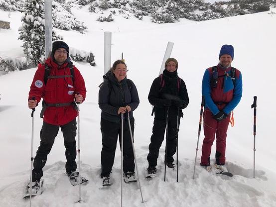 Four snowshoers with backpacks and long plexiglass tubes on their backs. All of them are wearing winter clothing and carrying hiking poles. Snow-covered mountain pines can be seen in the background. The person to the left has a bright red jacket with the logo of AWI (blue on white).