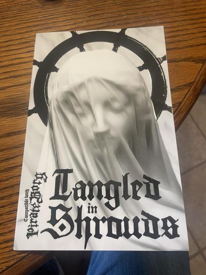 A black-and-white image of a booklet featuring a veiled figure, possibly a statue, with a halo-like design of a ship’s pilot wheel behind the head. The title "Tangled in Shrouds" is prominently displayed in bold, gothic lettering