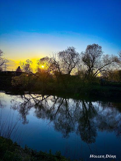 Ein ruhiger Frühlingsabend an der Lahn in Gießen. Die tief stehende Sonne wirft warmes, goldenes Licht durch die kahlen oder gerade austreibenden Äste hoher Bäume. Ihr Licht spiegelt sich sanft auf der glatten Wasseroberfläche des Flusses. Die Szene wirkt still, friedlich und leicht verträumt.