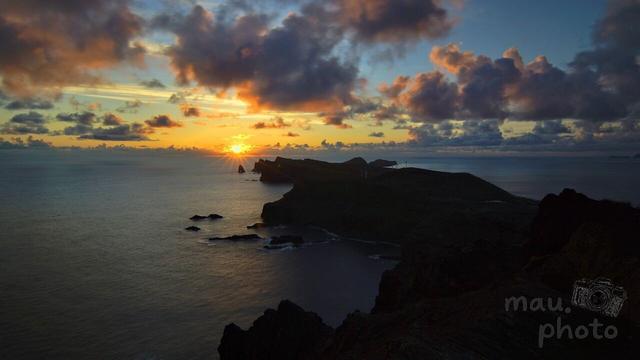 This image shows a scenic view of a coastline at sunset. The sky is filled with clouds, and the sun is partially obscured by the horizon, casting a warm glow over the ocean and the land. The dark silhouette of the coastline extends into the water, creating a striking contrast with the colorful sky and sea.