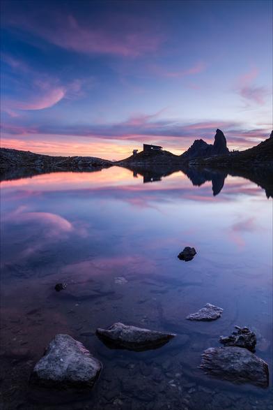 Photo d'un paysage de montagne au coucher du soleil au format portrait.
Au premier plan, quelques cailloux émergent près du bord d'un lac. Au bout du lac, un refuge et un pic rocheux en forme de canine se détachent sur fond de ciel rose et bleu. Dans le ciel, quelques fins nuages roses forment des virgules qui se reflètent dans le lac.
-
Portrait photo of a mountain landscape at sunset.
In the foreground, a few pebbles emerge near the edge of a lake. At the end of the lake, a refuge and a canine-shaped rock peak stand out against a pink and blue sky. In the sky, a few thin pink clouds form commas that are reflected in the lake.