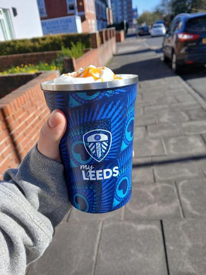 A white hand holding a blue leeds united travel cup. The lid is off, and you can see whipped cream with caramel on it