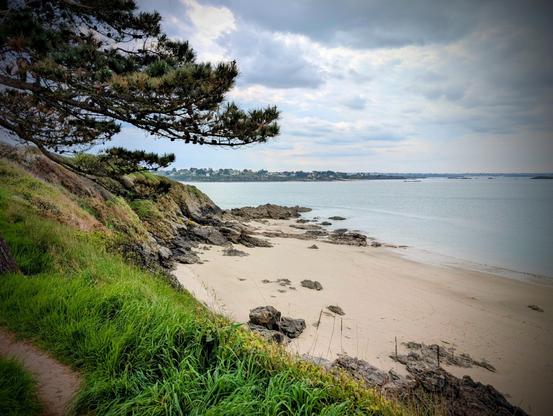 La côte avec l'océan, une plage, des rochers et un chemin herbeux. Le ciel est orageux mais encore beau.