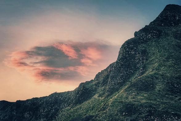 A wide, low-angle shot captures a rugged, dark green mountainside dominating the right side of the frame. The mountain's texture is rough, with visible ridges and patches of sparse vegetation. The sky above transitions from a light grayish-blue at the top to the soft, pale orange and pink of the sunset near the horizon on the left. A cluster of dark, pink-tinged clouds floats in the center-left of the sky, contrasting with the lighter hues. The silhouette of another lower mountain range is visible along the bottom left of the image.