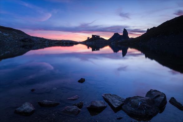 Photo d'un paysage de montagne au coucher du soleil.
Au premier plan, des cailloux baignent dans les eaux peu profondes d'un bord de lac. De l'autre côté du lac, un refuge se détache sur le ciel rose-orangé, juste à côté d'un pic montagneux dressé comme une canine. Ils se reflètent dans le lac.
-
Photo of a mountain landscape at sunset.
In the foreground, pebbles bathe in the shallow waters of a lakeshore. On the other side of the lake, a hut stands out against the pink-orange sky, right next to a canine-like mountain peak. They are reflected in the lake.