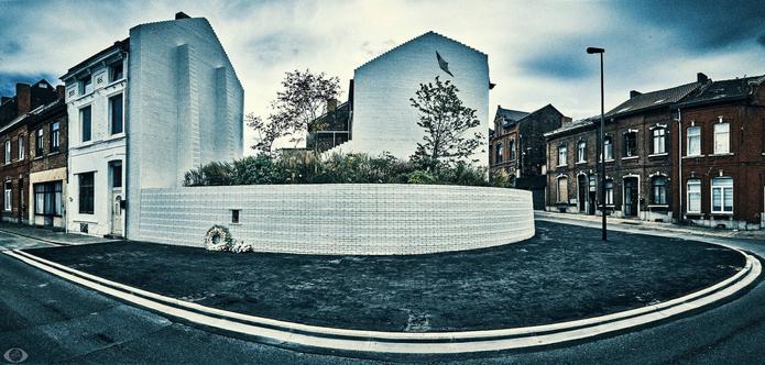 Low white wall that follows the lines of where the corner house of Dutroux stood. Behind it a lush garden. The walls that borders the houses on both sides are also whit. A flying bird is painted on the far wall. A commemorative wreath is places on the left just under a hall in the small wall where a flame is burning.

The atmosphere that the photo exhibits mimics that of the place.