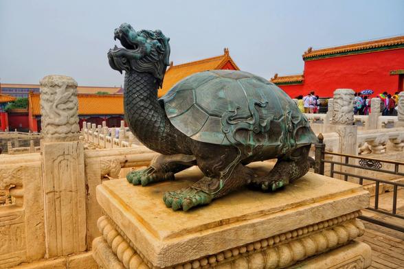 A detailed, eye-level shot captures a large, antique bronze statue of a mythical tortoise with a dragon's head, prominently displayed on an ornate stone pedestal. The creature's shell is textured with segmented patterns, and its powerful legs end in clawed feet. Its long, scaled neck rises to a fierce dragon head, complete with horns, whiskers, and an open mouth revealing teeth. The bronze has a weathered, dark green patina, suggesting its age.
The pedestal is made of light-coloured stone and features intricate carvings on its tiered base and top surface. The statue and pedestal are situated on a stone balustrade, adorned with carved pillars featuring cloud or scroll designs.
In the background, the vibrant colours and traditional architecture of what appears to be the Forbidden City in Beijing are visible. Buildings with bright red walls and golden-yellow tiled roofs stand under a slightly overcast sky. Distant figures of people, some holding umbrellas, suggest a tourist attraction.
