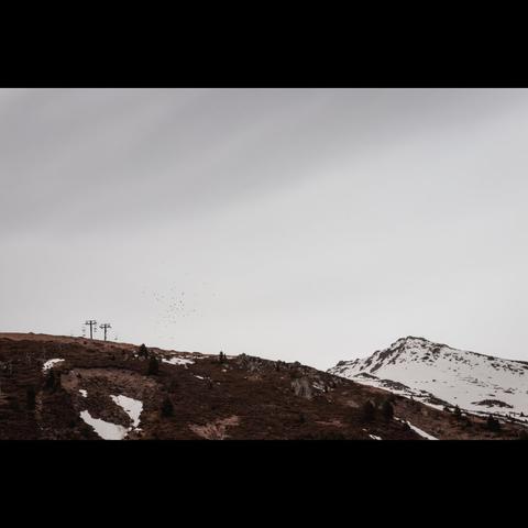 A landscape where you can see part of a mountain below, with some snow on the right. On the left, there's no snow, and two chairlifts can be seen. Above, you can see a large, cloudy sky.