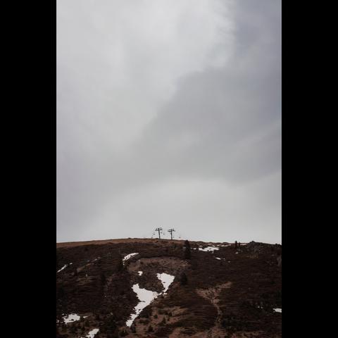 A vertical photography landscape where you can see part of a mountain below. On the center, there's no snow, and two chairlifts can be seen. Above, you can see a large, cloudy sky.