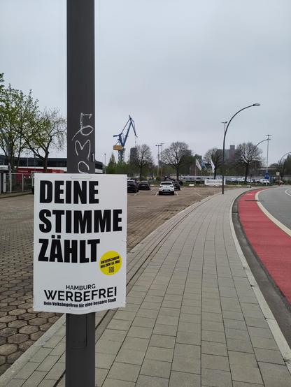 An einer recht neuen Straße, an einem Laternenmast: A1-Plakat mit "Deine Stimme zählt. Hamburg Werbefrei - unterschreib bis zum 13. Mai"