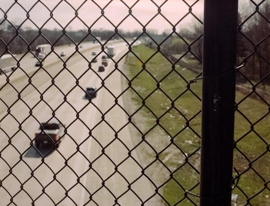 Cars and trucks on a highway as seen through a chainlink fence from the perspective of an overpass bridge.